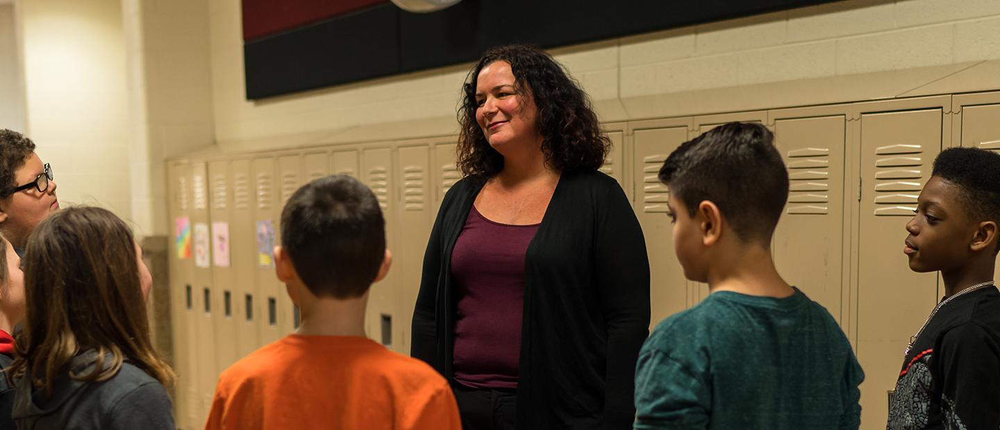 A woman standing in a hallway with lockers behind her and young students in front of her.